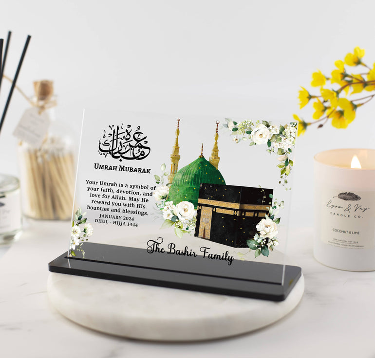 A clear acrylic plaque with a floral pattern and Islamic script, featuring a Kaaba art design, mounted on a black stand. It includes personalization details such as a family name and date.
