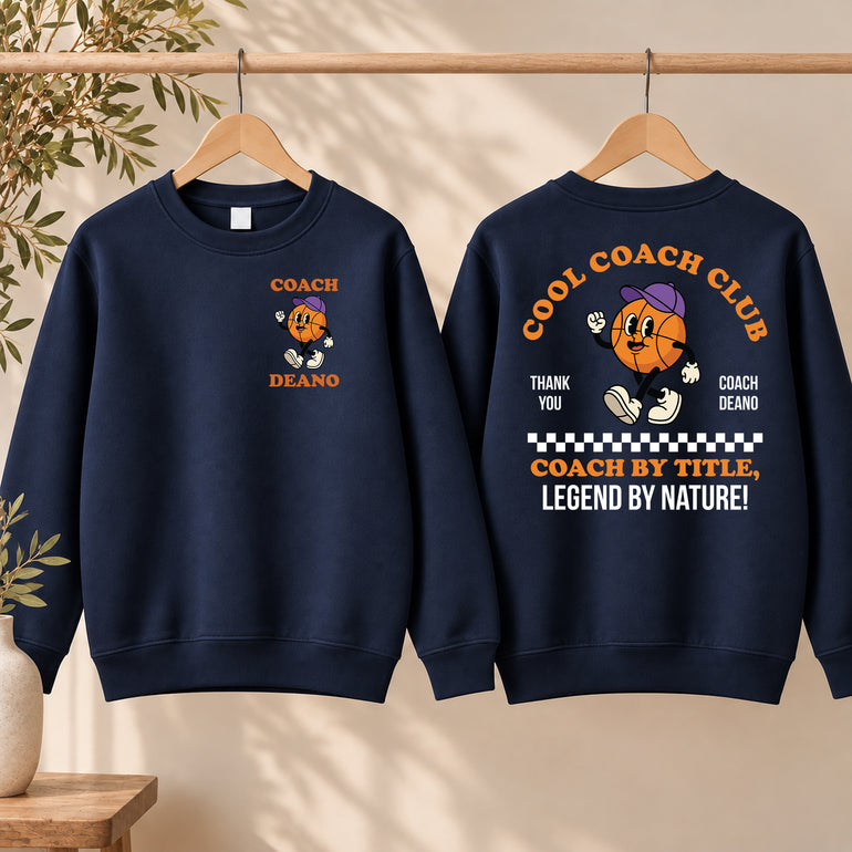 Navy Cool Coach Club basketball coach gift sweatshirt on a hanger, the back printed with Cool Coach Club, a retro cartoon basketball character in a purple cap, Thank You, Coach Deano and Coach by Title, Legend by Nature, the front with a small Coach Deano badge