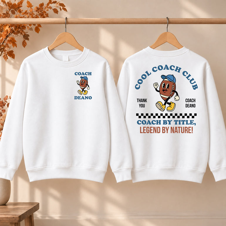 White Cool Coach Club rugby coach gift sweatshirt on a hanger, the back printed with Cool Coach Club, a retro cartoon rugby ball character in a blue cap, Thank You, Coach Deano and Coach by Title, Legend by Nature, the front with a small Coach Deano badge