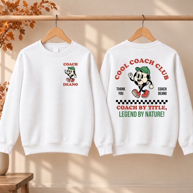 White Cool Coach Club football coach gift sweatshirt on a hanger, the back printed with Cool Coach Club, a retro cartoon football character in a green cap, Thank You, Coach Deano and Coach by Title, Legend by Nature, the front with a small Coach Deano badge