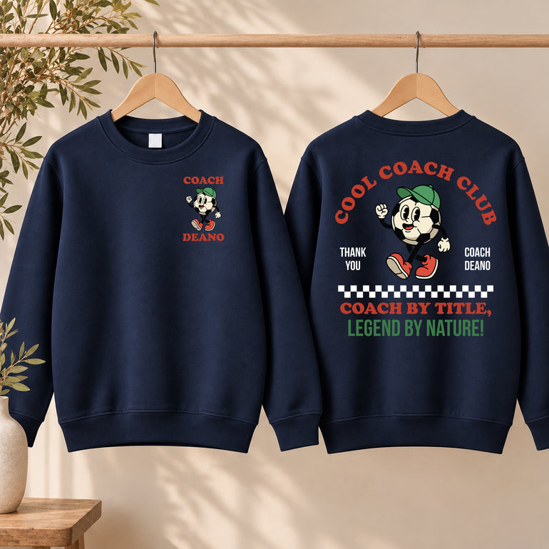 Navy Cool Coach Club football coach gift sweatshirt on a hanger, the back printed with Cool Coach Club, a retro cartoon football character in a green cap, Thank You, Coach Deano and Coach by Title, Legend by Nature, the front with a small Coach Deano badge