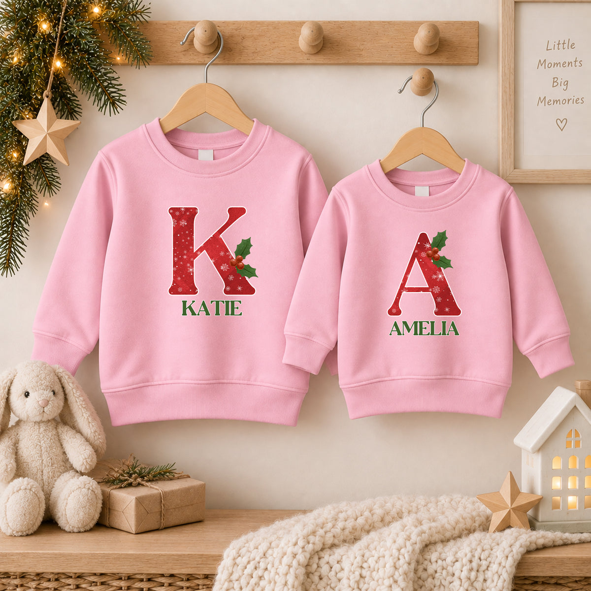Two light pink snowflake Christmas jumpers each with a large red snowflake initial, holly and the names Katie and Amelia in green