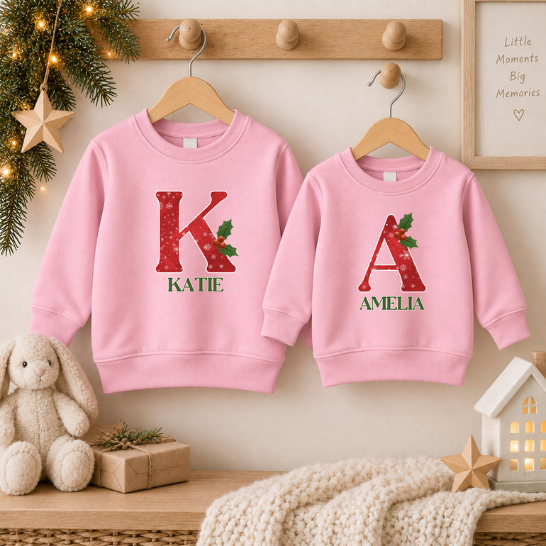 Two light pink snowflake Christmas jumpers each with a large red snowflake initial, holly and the names Katie and Amelia in green