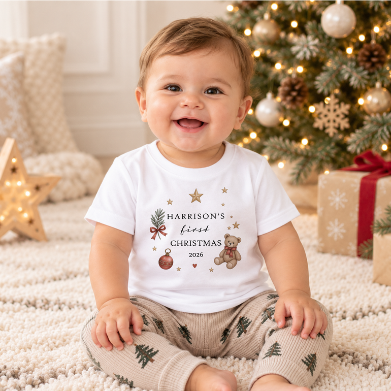 A smiling baby sitting on a white textured rug in front of a decorated Christmas tree, wearing a white graphic t-shirt that reads 'Harrison's first Christmas 2026' with a teddy bear and ornaments, and beige knit pants with green pine tree prints.