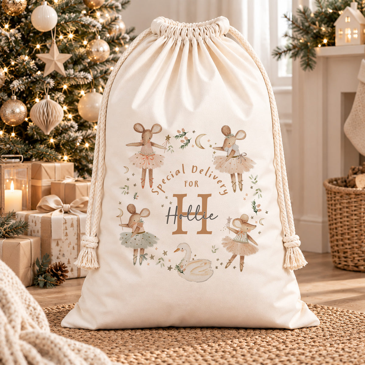 Cream personalised Christmas sack with ballerina mice, a swan and a child’s initial and name, beside a decorated Christmas tree.