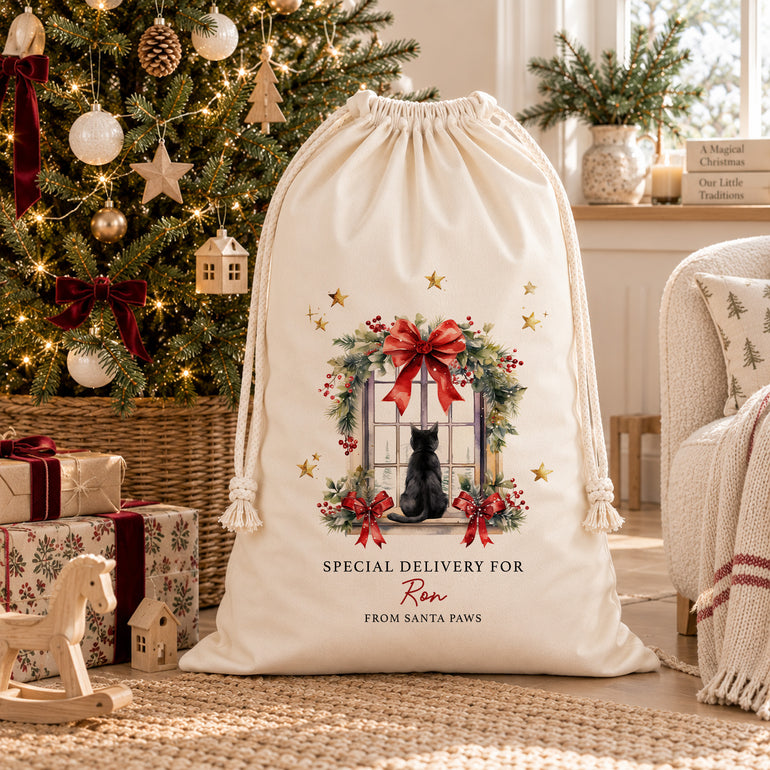 Cream personalised cat Christmas sack with a black cat at a festive window, displayed beside a decorated Christmas tree and wrapped presents.