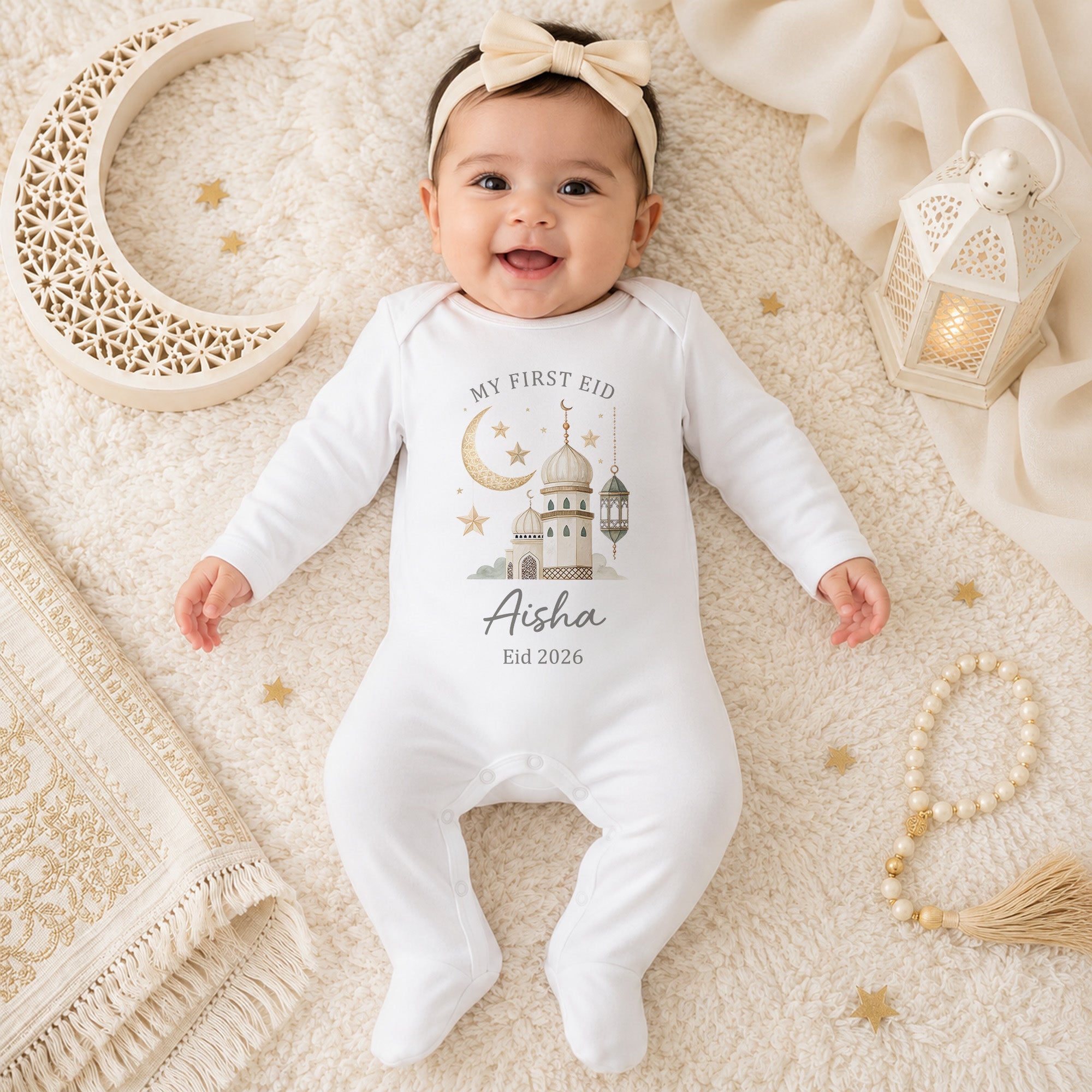 Baby wearing a neutral personalised First Eid baby grow with mosque, crescent moon, lantern and name design on a cream Eid background.