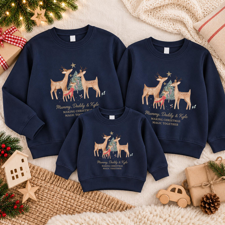 Three navy matching family Christmas jumpers laid flat, two adult and one child, each with a reindeer family around a Christmas tree, example family names in gold and Making Christmas Magic Together