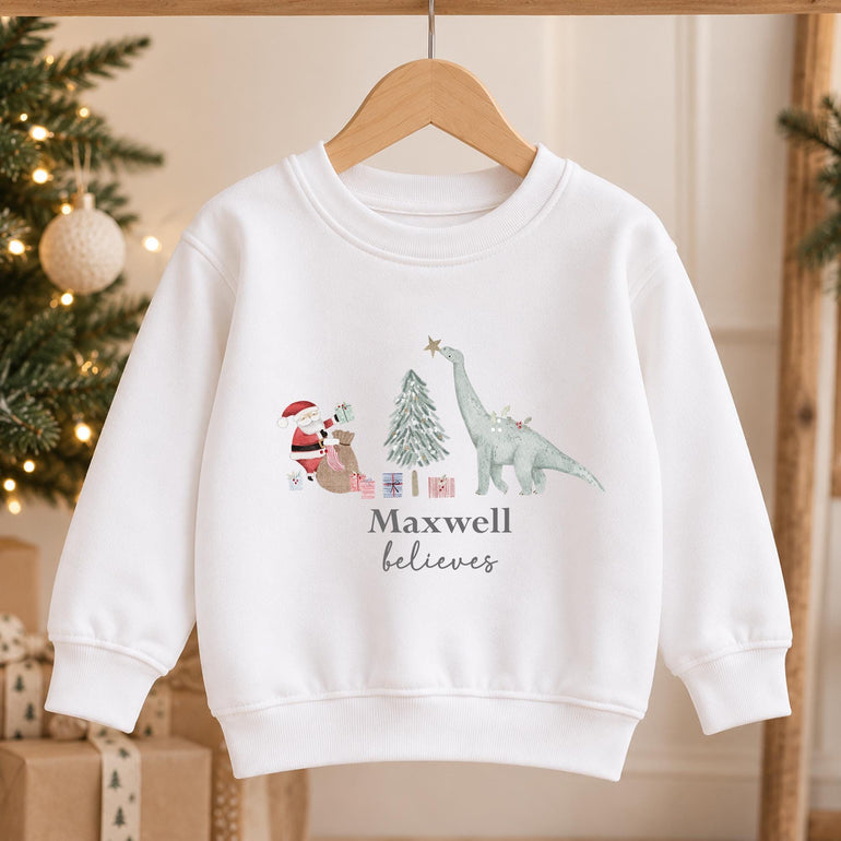 White kids dinosaur Christmas jumper on a hanger with the Santa and dinosaur design and a name in grey