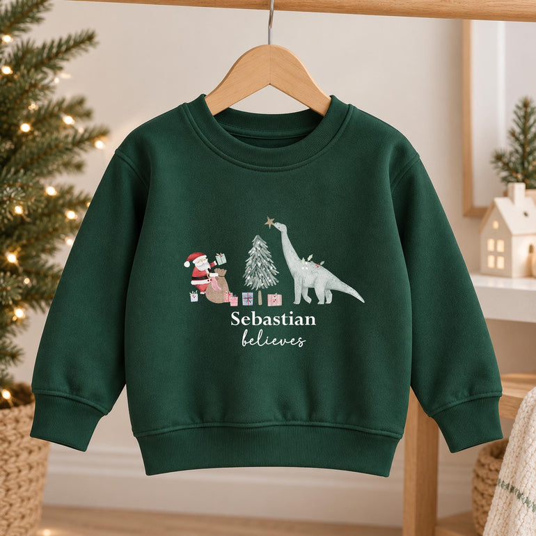 Forest green personalised dinosaur Christmas jumper on a hanger, with Santa, a Christmas tree and a long-necked dinosaur above a child's name and the word believes