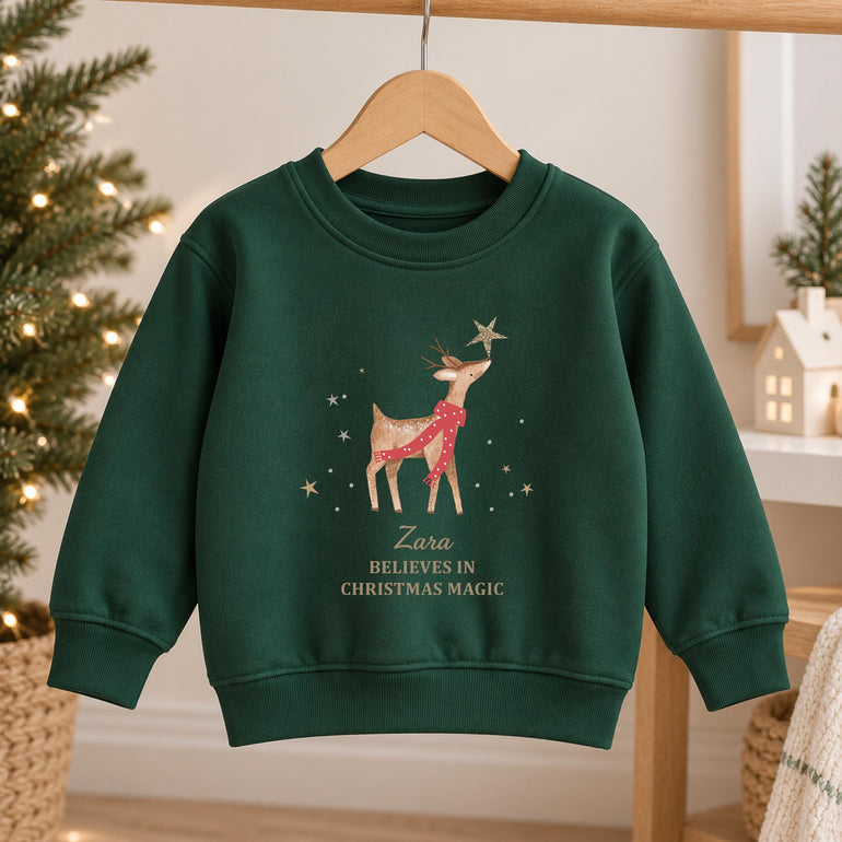 Forest green kids reindeer Christmas jumper on a hanger, with a reindeer in a red scarf touching a gold star above a name and Believes in Christmas Magic