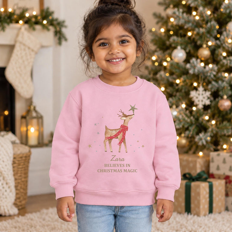 Young girl smiling beside a Christmas tree wearing the light pink personalised reindeer Christmas jumper