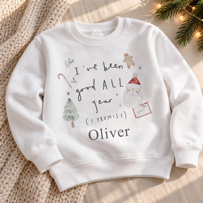 White kids Christmas jumper laid flat on a knitted blanket, with grey I've been good ALL year lettering, a Santa and letter illustration and a name
