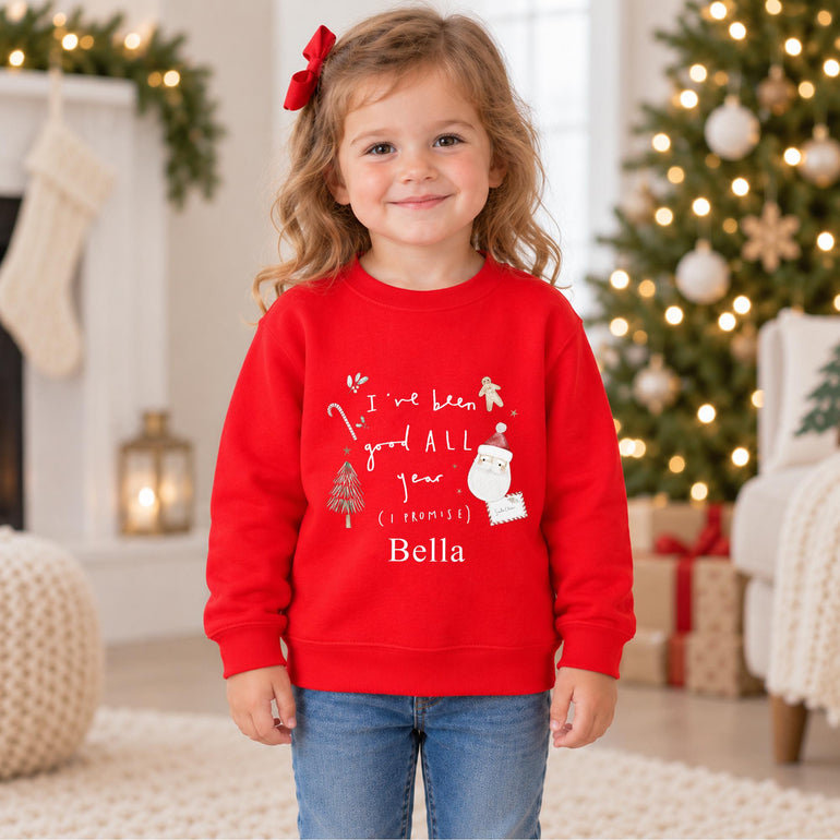 Young girl in front of a Christmas tree wearing a red personalised kids Christmas jumper reading I've been good ALL year (I promise) with her name