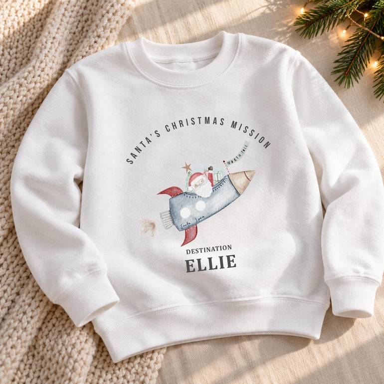 White personalised kids Christmas jumper laid flat, with the Santa rocket design and a name in grey lettering