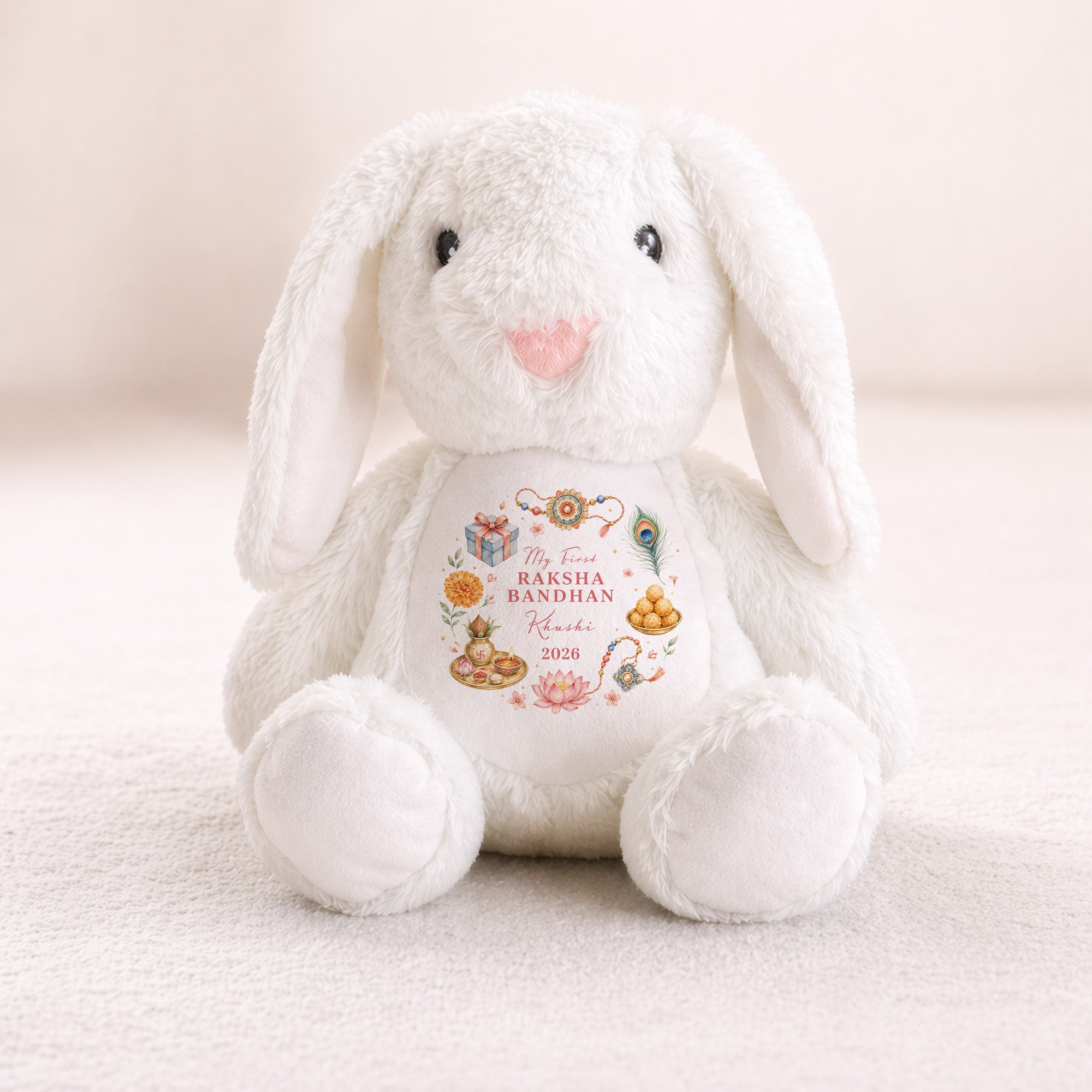 A white plush bunny toy with long floppy ears, black bead eyes, and a pink nose, sitting on a soft cream surface. The front of the bunny has a printed decorative label that reads 'My First RAKSHA BANDHAN 2026' with small colorful illustrations and personalised name