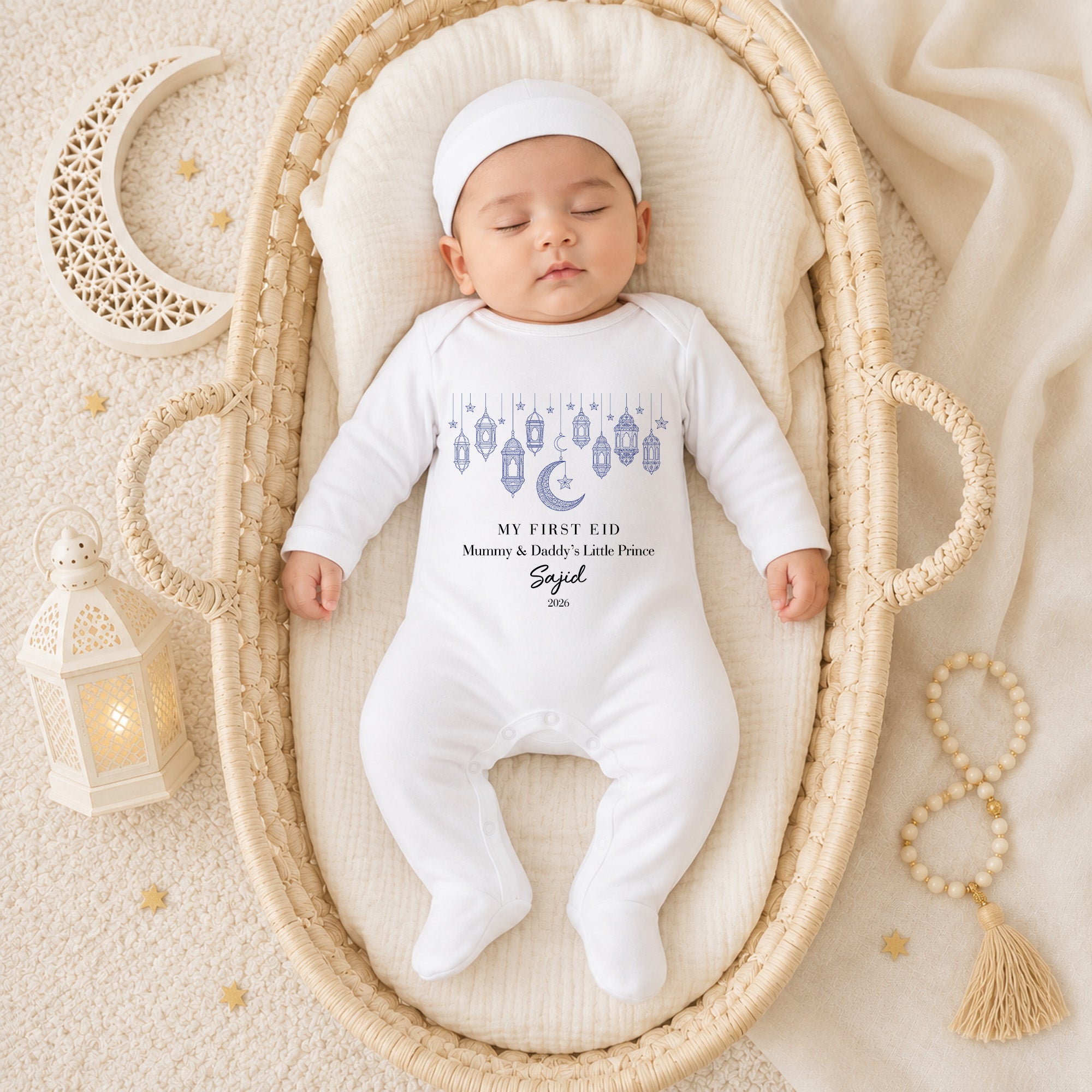 Sleeping baby wearing a personalised My First Eid baby grow with blue hanging lanterns, crescent moon and Little Prince wording in a wicker basket.