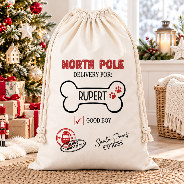 Personalised Santa Paws dog sack with red paw prints and festive postal stamps, displayed in a Christmas room beside wrapped gifts.